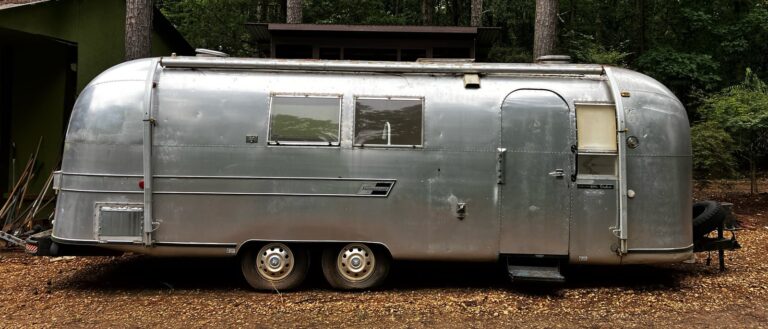 The PrayerStream: a 1967 Airstream travel trailer, seen side-on, parked on gravel among tall pines